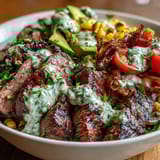A vibrant Tex-Mex bowl features tender steak, sliced avocado, and a generous drizzle of bright green cilantro cream sauce.